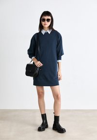 Σκούρο μπλε oversized φόρεμα με αντίθετο ανοιχτό μπλε γιακά, συνδυασμένο με μαύρες δερμάτινες μπότες μέχρι τον αστράγαλο και μαύρη τσάντα χιαστί.