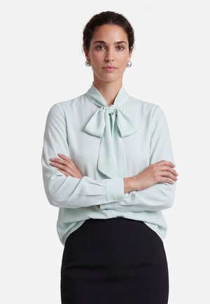Femme aux cheveux foncés attachés porte de grandes boucles d'oreilles en perles, un chemisier bleu clair avec un nœud et une jupe noire, les bras croisés avec assurance.
