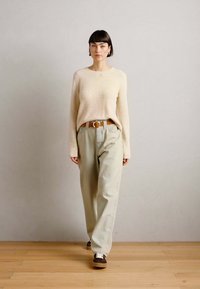 Beige textúrájú pulóver, laza világoszöld nadrág, barna öv és barna sportcipő fehér fűzősorral, áll a fából készült padlón, világos fal előtt.