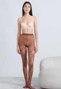 Καφέ καλσόν με δαντελένιο μοτίβο και διάφανο επάνω μέρος, αναδεικνύοντας ομαλή μετάβαση σε συμπαγές καφέ τμήμα στη μέση. Ανοιχτό μπεζ σουτιέν.
