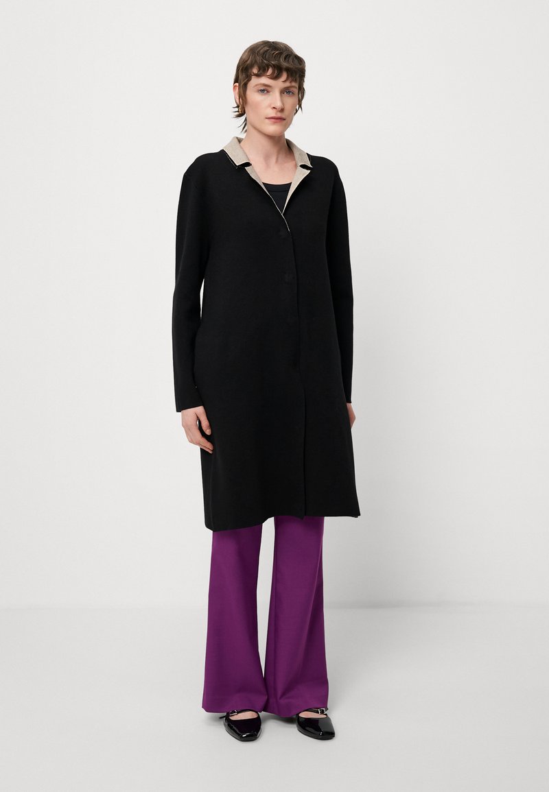 Cappotto lungo nero con colletto beige, chiusura a un pulsante, indossato sopra un top nero e pantaloni a gamba larga viola, abbinato a scarpe nere flat.