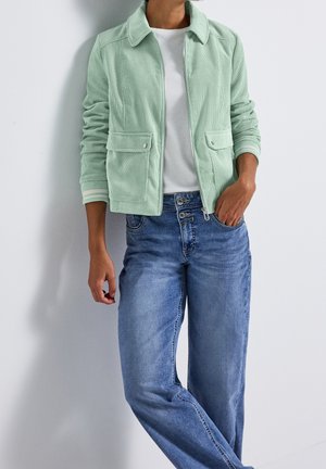 Osoba ubrana w jasnozieloną sztruksową kurtkę zapinaną na zamek, białą koszulę i niebieskie dżinsy z wysokim stanem, stojąca z jedną ręką w kieszeni na tle białej ściany.