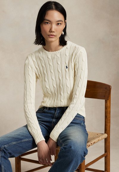 Jeune femme aux cheveux noirs portant un pull blanc torsadé et un jean bleu, assise sur une chaise en bois devant un fond neutre.