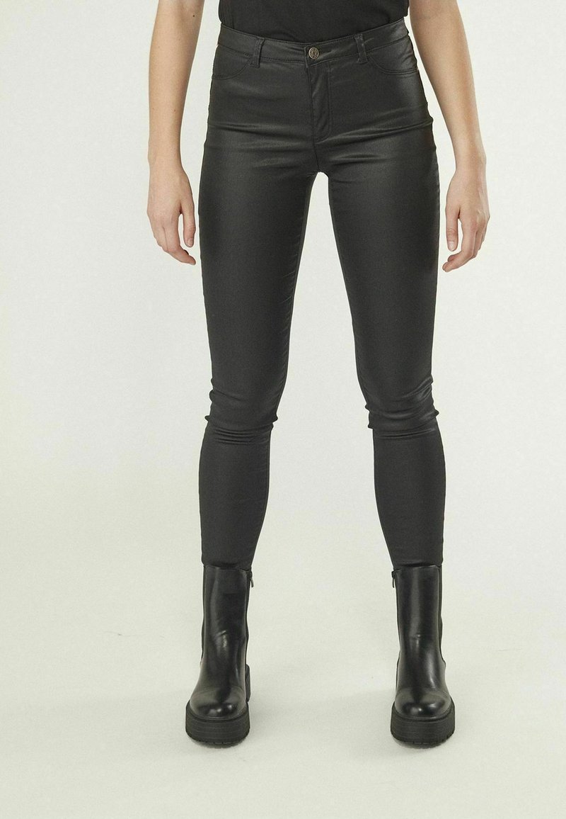 Pantalones ajustados de cuero sintético negros con una cinturilla alta y bolsillos delanteros, combinados con botas de tobillo negras de tacón grueso.
