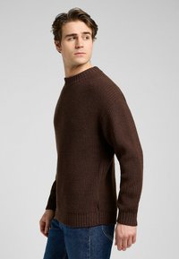 Brun stickad tröja med rund halsringning, texturerat tyg, ribbade ärmslut och nederkant samt sidofickor. Buren över blå jeans.