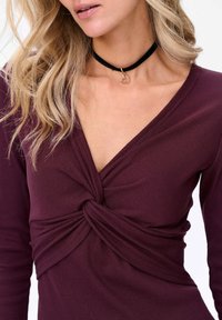 Bordowy prążkowany top z kręconym detalem z przodu i głębokim dekoltem w kształcie litery V. Połączony z czarnym naszyjnikiem, który ma złoty wisior w kształcie serca.