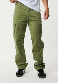 Pantaloni cargo verdi a taglio comodo, con multiple tasche laterali, realizzati in tessuto di cotone resistente, abbinati a scarpe da ginnastica nere.