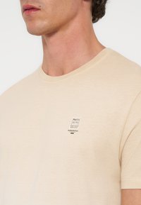 Άνδρας που φοράει ένα μπεζ t-shirt με στρογγυλή λαιμόκοψη και μικρό λογότυπο "Pier One Authentic Dept." στο αριστερό στήθος.