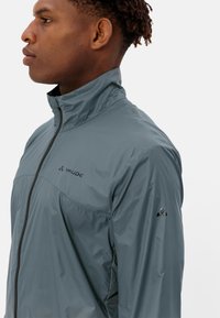 Leichte graue Jacke mit hohem Kragen, Frontreißverschluss und Logodetail auf der Brust und dem Ärmel, aus glattem, wasserabweisendem Material gefertigt.