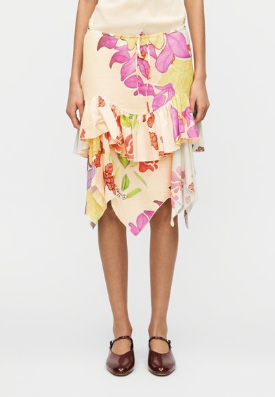 Jupe midi asymétrique à volants floraux dans des tons pastel portée avec des chaussures Mary Jane marron, montrant la taille et les jambes.