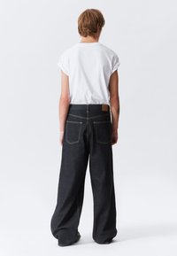 Persona con una camisa blanca de manga corta y jeans negros anchos y sueltos, de pie de espaldas contra un fondo claro y liso.