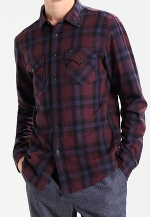 Hombre con una camisa de manga larga abotonada con un patrón de cuadros en rojo oscuro y azul marino, combinada con pantalones texturizados grises, manos en los bolsillos.