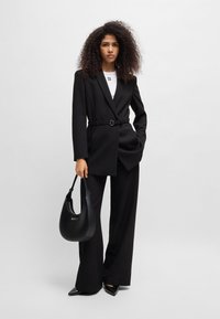 Černý tailored blazer s texturovanou látkou, páskem v pase, spárovaný s černými širokými kalhotami. Doplňky zahrnují černou ramenní tašku.