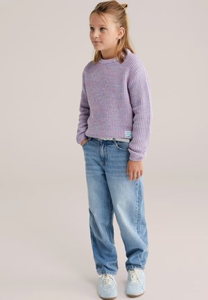 Enfant aux cheveux blonds portant un pull en tricot violet, un jean bleu ample et des baskets blanches, debout les mains dans les poches sur un fond uni.