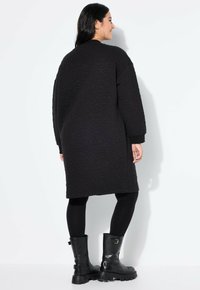 Abrigo largo de tejido acolchado negro con un ajuste relajado, que presenta puños acanalados y un cuello redondo, combinado con leggings negros y botas gruesas negras.