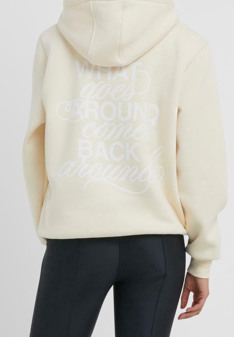 Personne portant un sweat à capuche crème avec un texte blanc « What goes around comes back around » dans le dos, associé à un pantalon noir.
