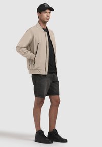 Beige bomberjakke med sorte detaljer, sort t-shirt, mørke denimshorts og sorte høj-top sneakers, præsenteret på en neutral baggrund.