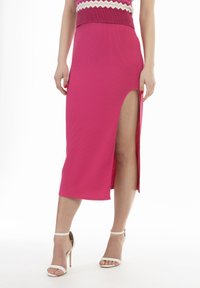 Gonna midi rosa a coste aderente con una spaccatura laterale, texture liscia e vita elasticizzata. Indossata con sandali bianchi con tacco alto e lacci.