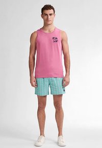 Růžové tílko bez rukávů s grafikou palmy, v kombinaci s krátkými kalhotami s tyrkysovými a bílými pruhy. Model stojí na světle šedém pozadí.