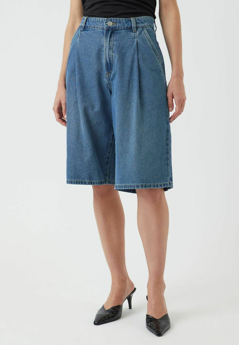Denim Bermuda shorts dengan lipatan, cuci biru, pinggang sedang, saku samping, dan hem kasar. Dipadukan dengan mules bertumit hitam.