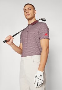 Hnědočervený kostkovaný polo trik s hnědým límečkem, zkombinovaný se světle béžovými kalhotami. Golfista drží hůl a má na sobě bílou rukavici s černými akcenty.