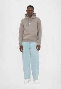 Felpa grigia con logo, tasca frontale e cordini, abbinata a jeans baggy azzurri e scarpe da ginnastica bianche. Design semplice e casual.