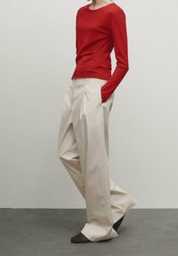 Haut rouge à manches longues associé à un pantalon beige taille haute à plis. Tissu lisse, coupe décontractée, et chaussures foncées à bout pointu.