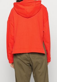 Sudadera corta roja de algodón con capucha con cordón, mangas largas y puños acanalados. Combinada con pantalones verde oliva.