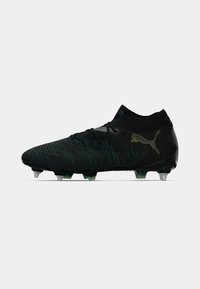 Botas de fútbol negras con una parte superior texturizada y franjas verdes, diseño de corte bajo y tacos moldeados para tracción. Acentuadas con un logo dorado.