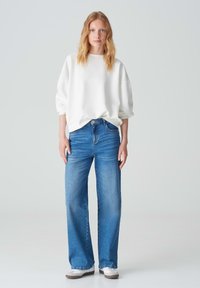 Felpa bianca oversize con spalle cadenti, abbinata a jeans a vita alta blu con gamba larga. Sneaker bianco e grigio completano l'outfit.