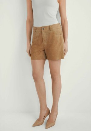 Kobieta w beżowych zamszowych szortach, białym topie bez rękawów i cielistych szpilkach z ostrym noskiem, stojąca na tle jasnego, jednolitego tła.