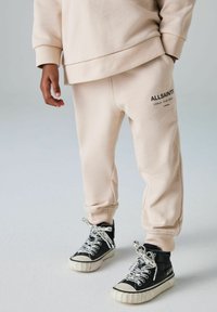 Beige Baumwoll-Sweatpants mit elastischen Bündchen und Seitentaschen, versehen mit einem schwarzen "ALLSAINTS"-Logo am linken Oberschenkel. Schwarze High-Top-Sneaker mit gemusterten Schnürsenkeln.