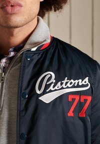 Giacca bomber navy con polsini grigi, caratterizzata da un logo ricamato "Pistons" in bianco e rosso, chiusure a scatto e un design sportivo.