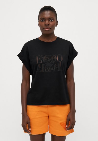 Tricou negru din bumbac cu mâneci rulou, având un logo metalic central „Emporio Armani”. Asortat cu șorturi portocalii cu detalii texturate.