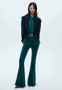 Marineblauwe cropped jas, teal knoopshirt en flared teal broek. Glanzende zwarte puntige schoenen. Glad textuur, gelaagd outfitontwerp.
