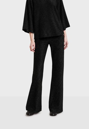 Donna che indossa pantaloni neri a gamba larga e un top largo abbinato con un motivo testurizzato sottile, in piedi davanti a uno sfondo chiaro e uniforme.