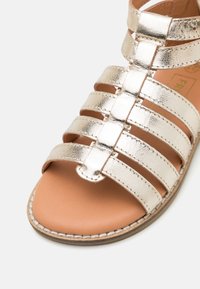Sandale mit offenen Zehen, mehreren metallic-goldenen Lederriemen und einer tanfarbenen gepolsterten Fußsohle mit sichtbaren Nähten entlang der Sohle.