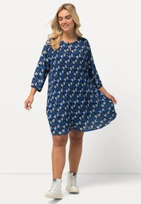 Nainen, jolla on pitkät vaaleat hiukset, pukeutuneena siniseen kukkakuosiseseen laskostettuun mekkoon ja valkoisiin korkean varren tennareihin, pitää mekon helmaa kädessään hymyillen.