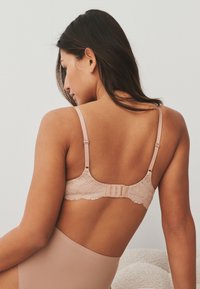 Frau mit dunklen Haaren, die seitlich sitzt und einen hautfarbenen Spitzen-BH sowie passende High-Waist-Unterwäsche vor einem schlichten Hintergrund trägt.