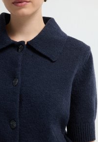 Marineblauer Cardigan aus strukturiertem Strickstoff, mit großem Kragen, schwarzen Knöpfen und gerippten Ärmeldetails.