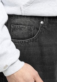 Jeans negre din denim cu un buzunar mic, etichetă "EIGHTYFIVE" și detalii cu buton argintiu, asortate cu manșonul unui hanorac gri.