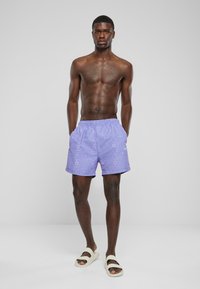 Shorts de bain violet à motifs avec une taille élastique, des poches latérales et une coupe décontractée, portés avec des sandales beiges sur des pieds nus.