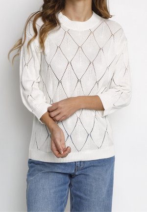 Femme portant un pull en maille blanc à motif losanges avec manches longues et un jean bleu, debout, les mains jointes devant elle.