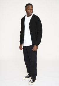 Sort ribbet cardigan med knapper, båret over en hvid t-shirt, kombineret med sorte bukser og lavtoppede sorte sneakers med hvide såler.