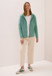 Felpa verde menta con zip e coulisse, due tasche frontali, abbinata a una maglietta bianca e pantaloni a gamba larga color crema.