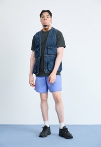Denim vest with multiple pockets, black t-shirt, light purple shorts, and black sneakers. Set against a plain background.