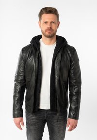 Giacca di pelle nera con cappuccio, dal design aderente, chiusura con zip e tasche laterali, indossata sopra una camicia bianca e jeans scuri.
