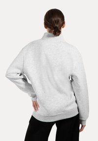 Persona con cabello castaño recogido en una baja coleta, vistiendo un sudadero gris claro y pantalones negros, de pie con la mano en la cadera, de espaldas al espectador.