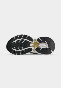 Semelle de chaussure noire et blanche avec des accents dorés, présentant des motifs de grip texturés et de petits clous ronds pour la traction.