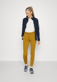 Jūrininkų zip-up striukė, smėlio spalvos marškinėliai, garstyčių spalvos kelnės, ir pilki sportbačiai. Minkšta medžiaga, prigludęs dizainas, su raišteliu juosmenyje ir užtrauktuku užsegamu kišenėmis.